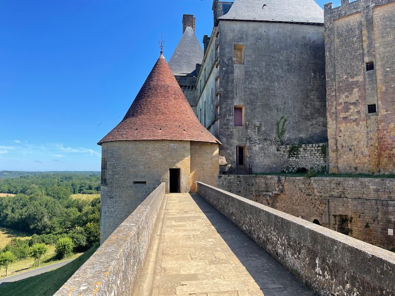 Een stenen pad naar een ronde toren van Château de Biron, met een bebost landschap op de achtergrond