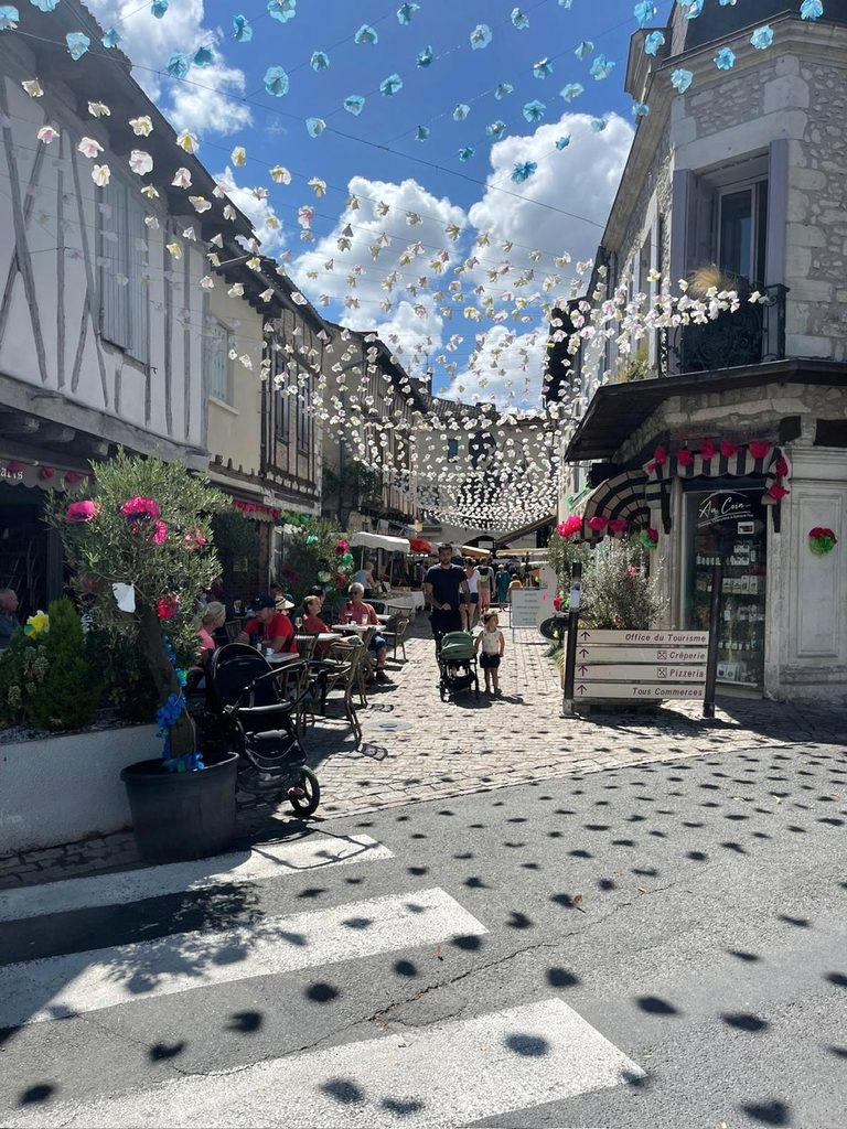 Caféterrasjes en vakwerkhuizen onder hangende versieringen in een geplaveide straat in Eymet
