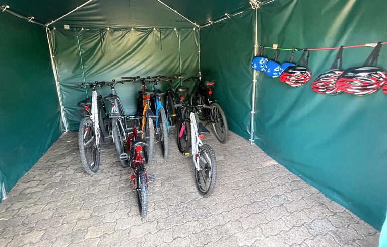 Erwachsenenräder und kleinere Fahrräder im überdachten Fahrradbereich der Maison Sérénité, daneben aufgehängte Helme
