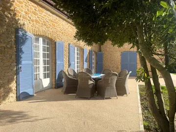 Round outdoor dining table and woven chairs beneath a tree beside the stone house
