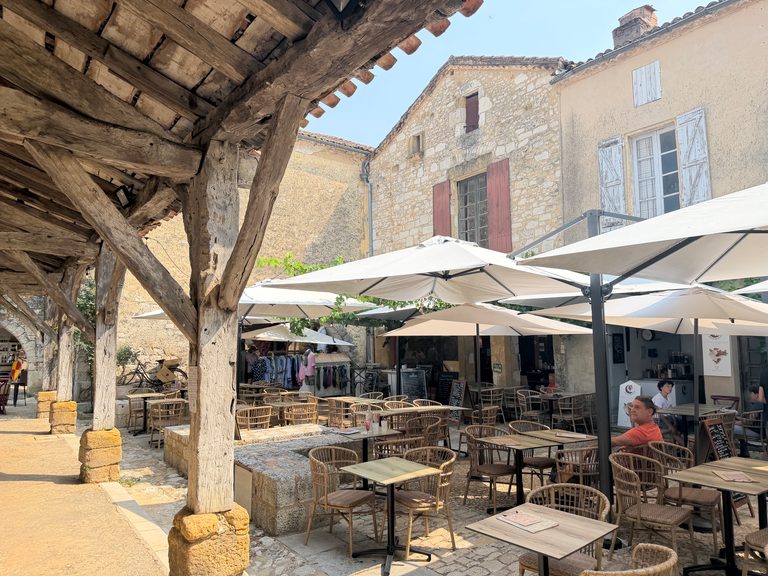 Cafétafels onder parasols naast stenen arcaden met houten steunpilaren op het plein in Monpazier